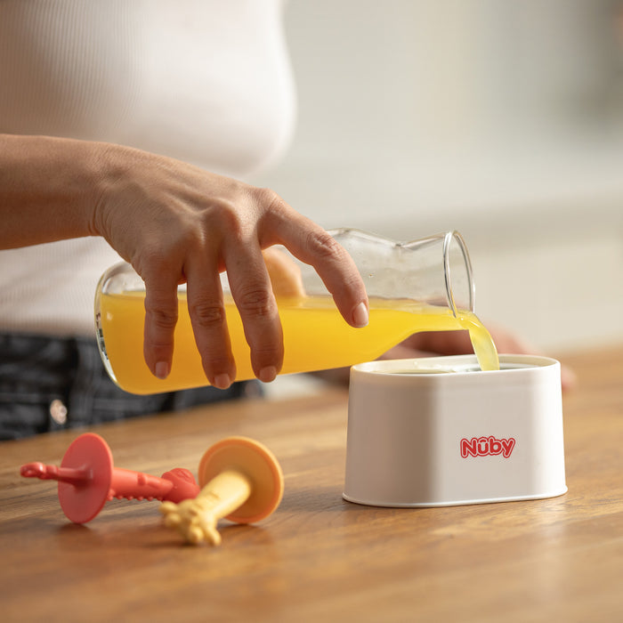 A person pours orange juice from a glass carafe into a white Nuby container on a wooden table, with two red and yellow Nuby Animal Pops Ice Pop Mold and homemade popsicles lying nearby.