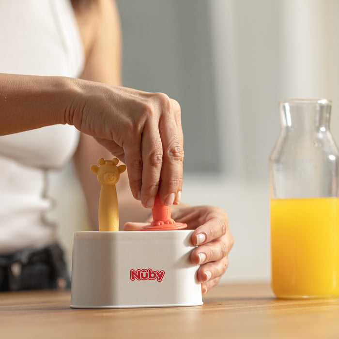 Someone uses the Nuby Animal Pops Ice Pop Mold, pressing it into a white container on a table beside a bottle of orange juice—great for making homemade popsicles.
