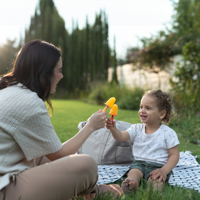 A woman and child sit on a blanket in the yard, smiling and clinking homemade popsicles made with the Nuby Animal Pops Ice Pop Mold as they enjoy a sunny outdoor day.