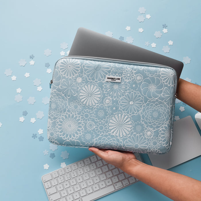 Hands holding a baby blue floral laptop sleeve with a laptop inside, next to a white keyboard on a blue surface.
