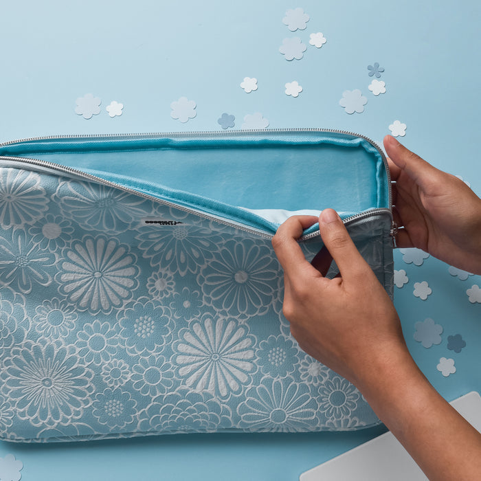 Hands opening a baby blue floral laptop sleeve on a blue surface, surrounded by paper flower cutouts.
