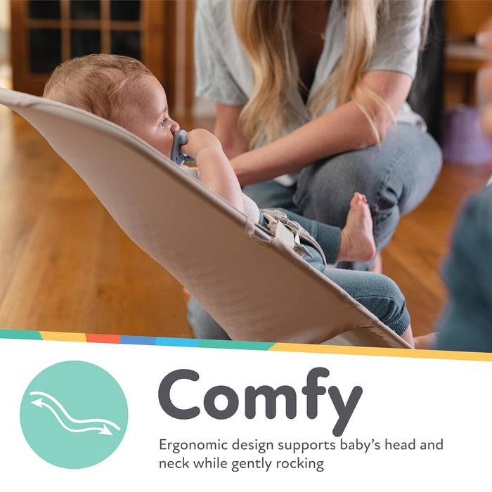 A baby relaxes in the Baby Bouncer Seat | Grey, sucking on a pacifier, while an adult kneels nearby. Text reads: Ergonomic design supports baby’s head and neck and gently rocks for soothing comfort.