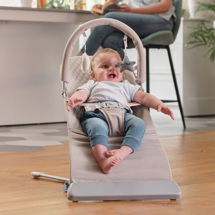 A smiling baby in a white shirt and blue pants sits indoors on a Baby Bouncer Seat | Grey with soft hanging toys, on a wooden floor. An adult is in the background near the kitchen counter.
