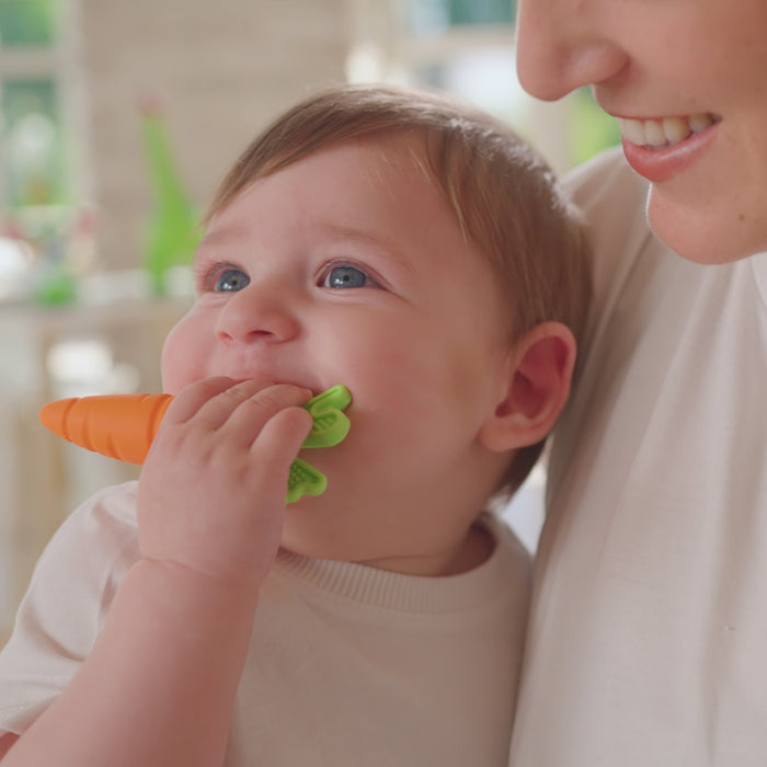 A smiling baby, dressed in white and held by an adult, plays with a Nuby Carrot Teether from the Carrot Teether Set (2 Pack) in a brightly lit room.