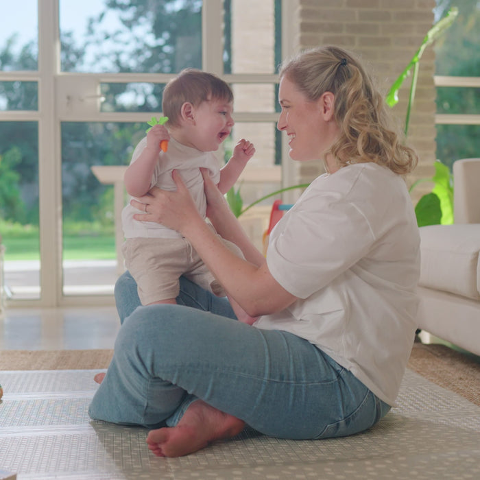 A woman sits cross-legged in a sunlit living room, lifting a smiling baby who holds the Nuby Carrot Teether Set (2 Pack), with large windows and green plants in the background.