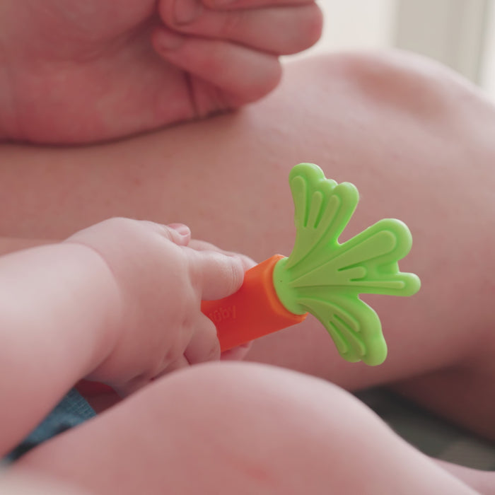 A baby holds a green and orange Nuby Carrot Teether from the Carrot Teether Set (2 Pack), with an adult's arm and hand visible in the background.