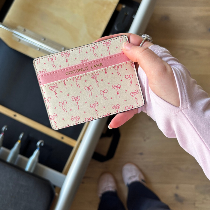 Hand holding a pink vegan leather Coconut Lane Dainty Bows Card Holder over a wooden floor with workout gear nearby.