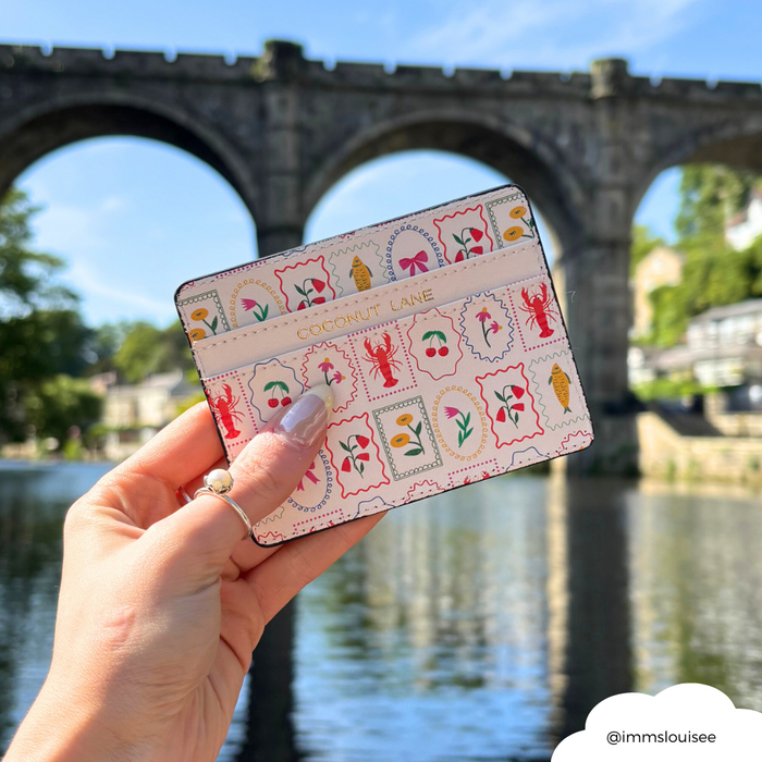 Hand holding Coconut Lane Dolce Vita card holder by a river, with stone bridge and trees in the background.