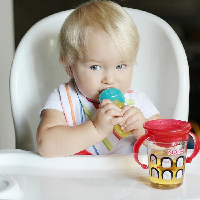A young blonde toddler in a high chair practices self-feeding with the E-Z Squee-Z Silicone Baby Food Dispenser in blue, wearing a striped bib, while a penguin sippy cup of juice sits nearby.