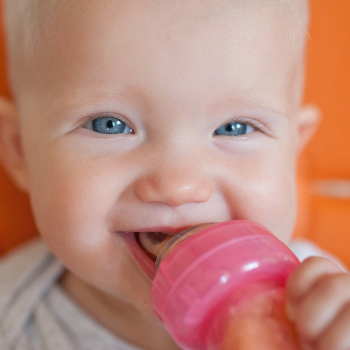 A smiling blue-eyed baby holds and chews on the E-Z Squee-Z Silicone Baby Food Dispenser in blue, perfect for self-feeding semisolid foods, with an orange background.