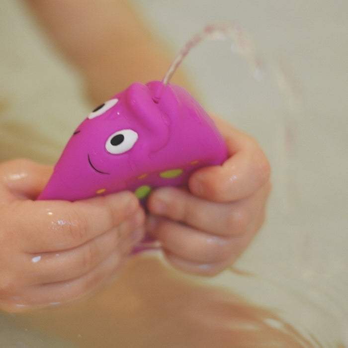A child’s hands hold a purple Fun Fish Squirter bath toy by Nuby, shaped like a whale, as it squirts water while partially submerged. Available in a 3-pack.