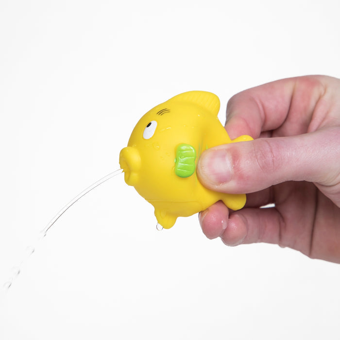 A hand squeezes a yellow Nuby Fun Fish Squirters bath toy from the 3-pack, making water squirt from its mouth against a white background.
