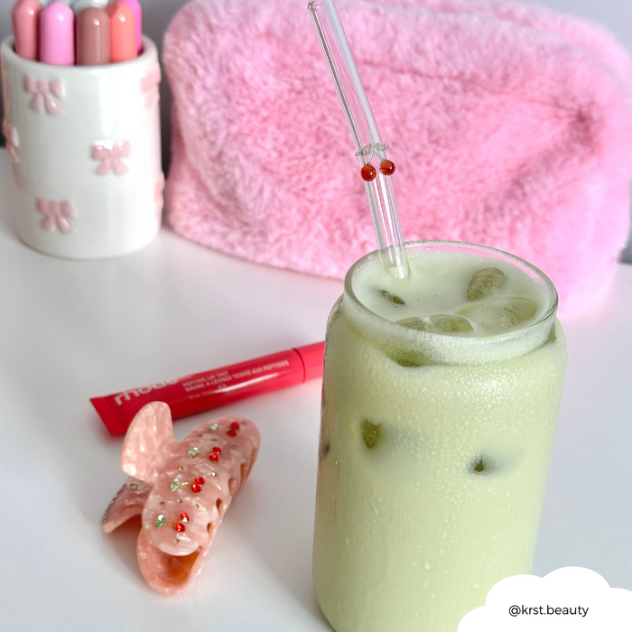 Glass of iced matcha latte with a clear glass straw featuring cherry and pink accents, next to a fuzzy pink pouch.