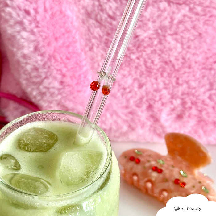 Iced green drink with Coconut Lane cherry-patterned glass straw, pink background, and a pink jeweled hair clip nearby.