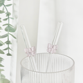 Pink Coconut Lane glass straw with a bow in a ribbed clear glass, next to green leaves and a sheer white curtain.