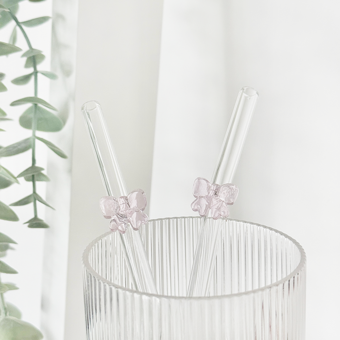 Pink Coconut Lane glass straw with a bow in a ribbed clear glass, next to green leaves and a sheer white curtain.