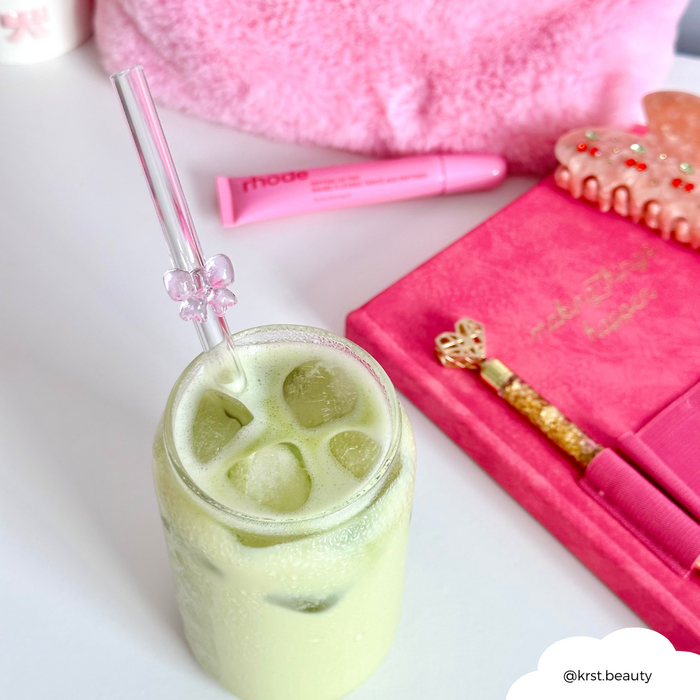 Iced matcha latte with a pink-bowed glass straw, next to pink beauty products, a notebook, and a gold pen.
