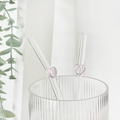 Two pink heart glass straws in a ribbed glass with green leaves blurred in the background.