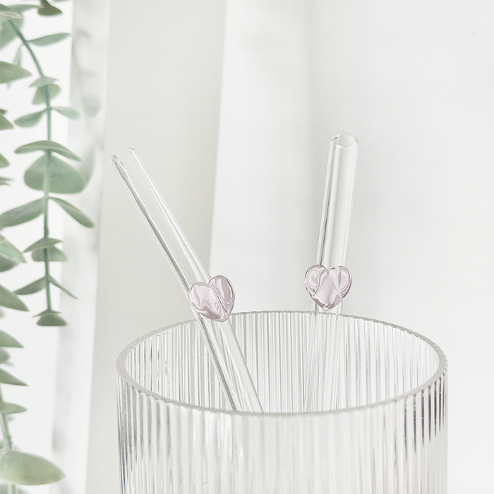 Two pink heart glass straws in a ribbed glass with green leaves blurred in the background.