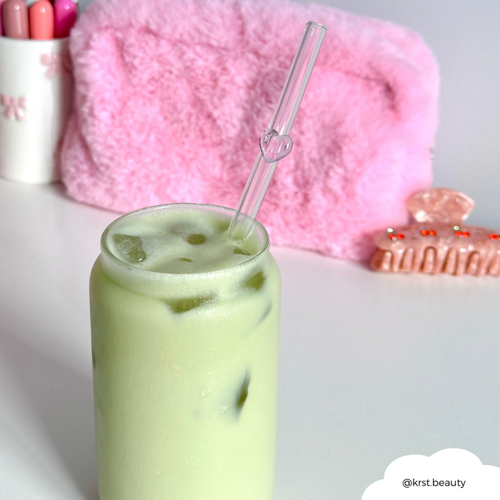 Iced matcha latte with a pink heart glass straw, next to a pouch, pink hair clip, and lipsticks.