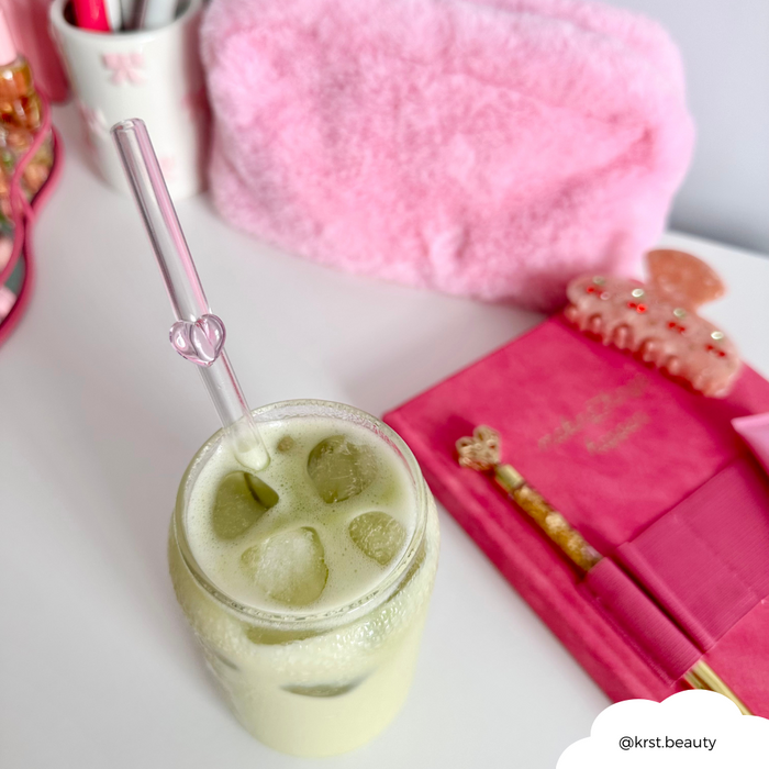Iced matcha latte with a pink heart glass straw in a clear cup on a pink desk with notebook and accessories.