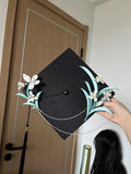 A black graduation cap adorned with white handmade Chanhua and turquoise - colored leaves, held by a hand