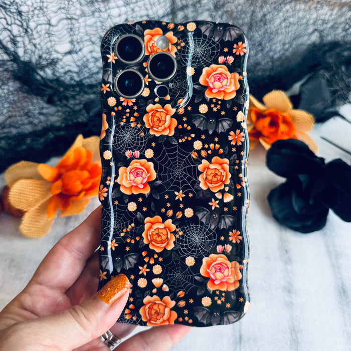 Haunted Blossoms Wavy Luxe MagSafe iPhone case held in hand, black with coral roses, gold spiderwebs and tiny bats.