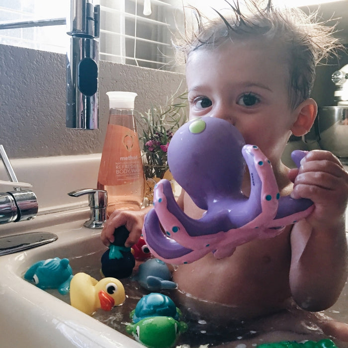 A young child with wet, spiky hair plays in a sink bath, holding a purple octopus from the Little Squirts Bath Squirters 10 pack. Colorful bath toys and rubber duckies float nearby as a bottle of body wash sits by the faucet.