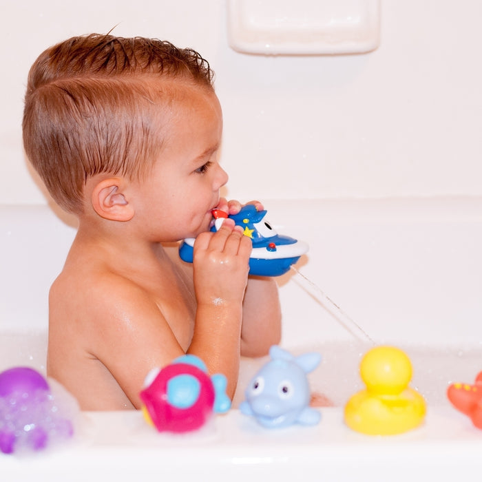 A young child with wet hair sits in a bathtub, playing with Little Squirts Bath Squirters—a 10-pack of colorful animal-shaped bath toys, including a duck and dolphin.