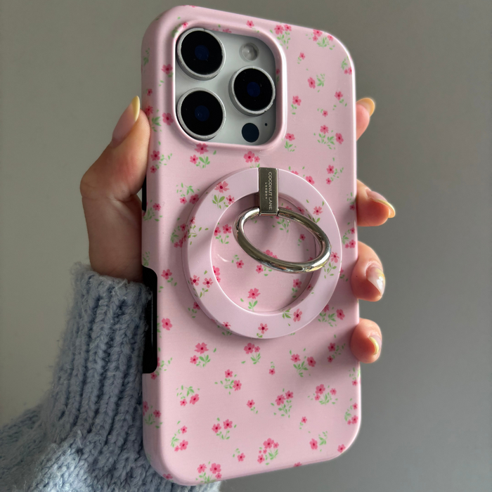 Hand holding iPhone with Coconut Lane MAGSAFE phone ring in pink ditzy floral pattern against a plain background.