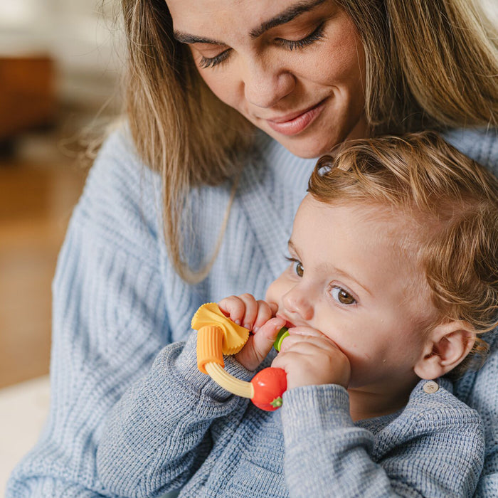 A woman smiles lovingly while holding a toddler who is chewing on a colorful silicone teething toy. Both are wearing light blue sweaters and sitting close together indoors.