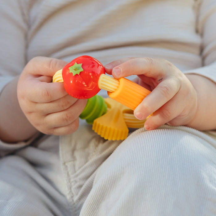 A baby in light-colored clothes holds the Nuby Pasta Silicone Teething Ring, a colorful silicone teether with a red tomato shape and yellow-orange handles.