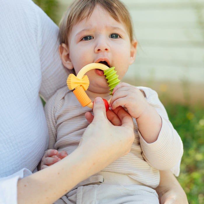 A baby sits on an adult’s lap outdoors, chewing on the colorful Nuby Pasta Silicone Teething Ring while the adult supports them, looking slightly uncomfortable.