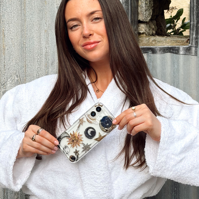 Woman in a white robe smiles, holding a clear phone case with a celestial print near a rustic wall.