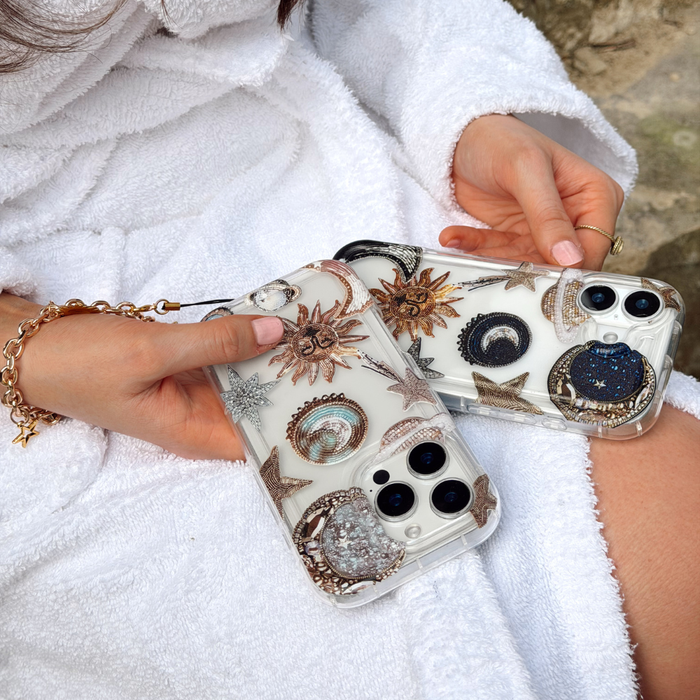 Hands holding Coconut Lane clear phone case with celestial print; wearer has a white robe and gold bracelet.