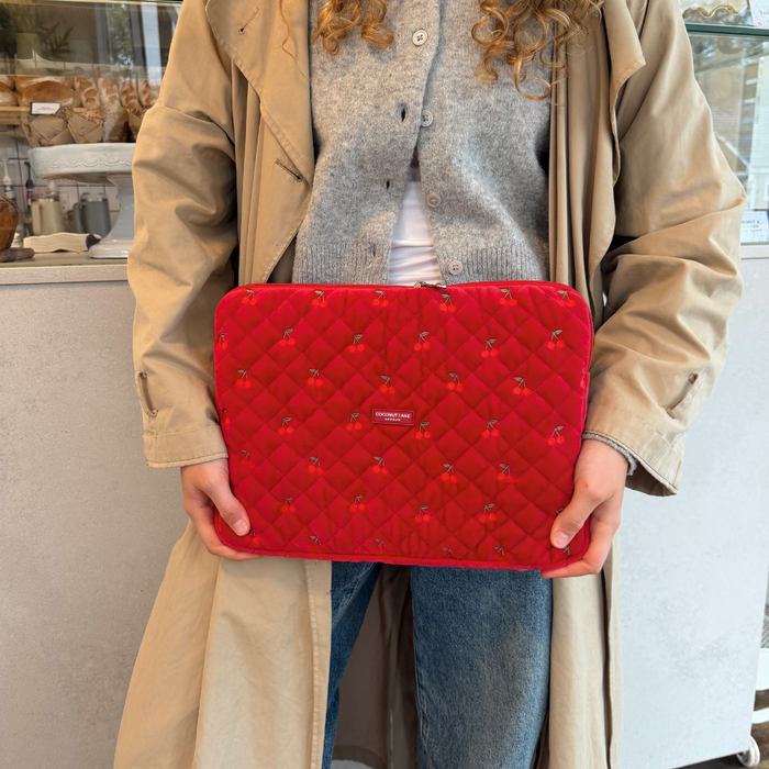 Tan coat person holds a red quilted velvet laptop sleeve with cherry pattern beside a pastry counter.