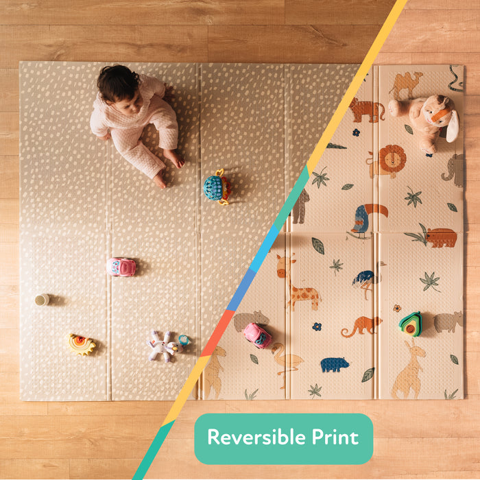 A baby sits on a light wooden floor atop the Reversible Floor Mat | Safari, which features two designs: a beige dotted pattern on one side and a safari animal print on the other. A label reads "Reversible Print.