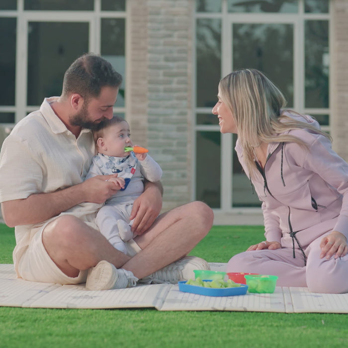 A man and woman sit on a picnic blanket outdoors, smiling at each other while the man soothes their baby with the Nuby Sensory Carrot Teether. Colorful containers of food are spread out on the blanket.