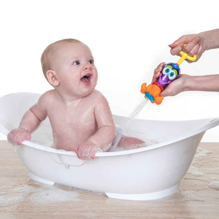 A smiling baby sits in a white bathtub while an adult playfully sprays water with the Squid Squirter Water Squirter, making bath time fun and interactive as the giggling baby enjoys the soapy bubbles.