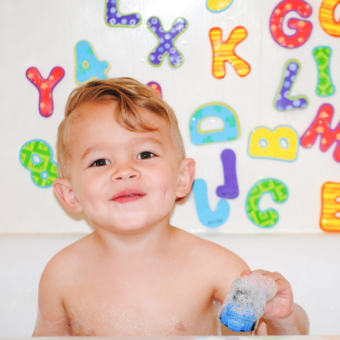 A smiling young child sits in the bathtub holding a Nuby Squirt Wheels Bath Squirters - 4 pack, with colorful foam alphabet letters decorating the white tiled wall behind, making bath time fun and full of imaginative play.