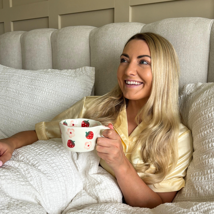Woman with long blonde hair in bed holds a white mug with pink strawberries and floral pattern from Coconut Lane.