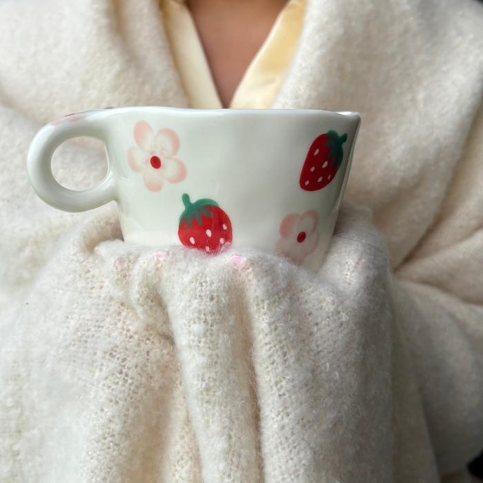 Person in a cream blanket holding a white mug with pink strawberries and floral pattern from Coconut Lane.