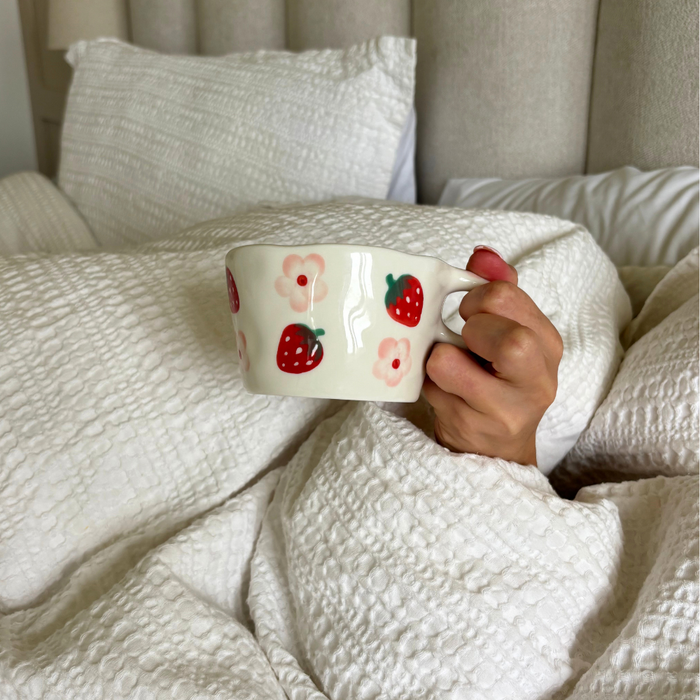 Hand holding Coconut Lane Strawberries & Flowers ceramic mug with pink and floral pattern on white bedding.