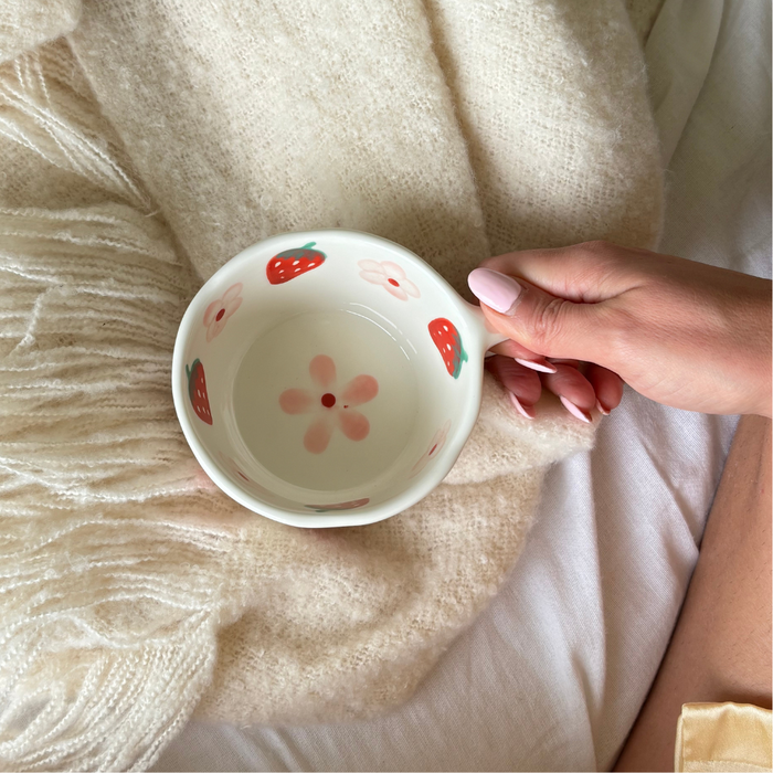 Hand with pink nails holding Coconut Lane Strawberries & Flowers ceramic mug over a soft, light-colored blanket.
