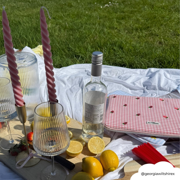 Coconut Lane Strawberry Picnic laptop sleeve with a pink pattern on grass, surrounded by wine, glasses, lemons, and candles.