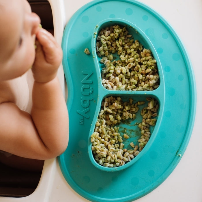 A baby sits in a high chair, eating with their hands from the Sure Grip Miracle Mat 2-Section Plate in grey, filled with green and white grains. The focus is on the suction plate and baby's hand, with the baby's face partially visible.