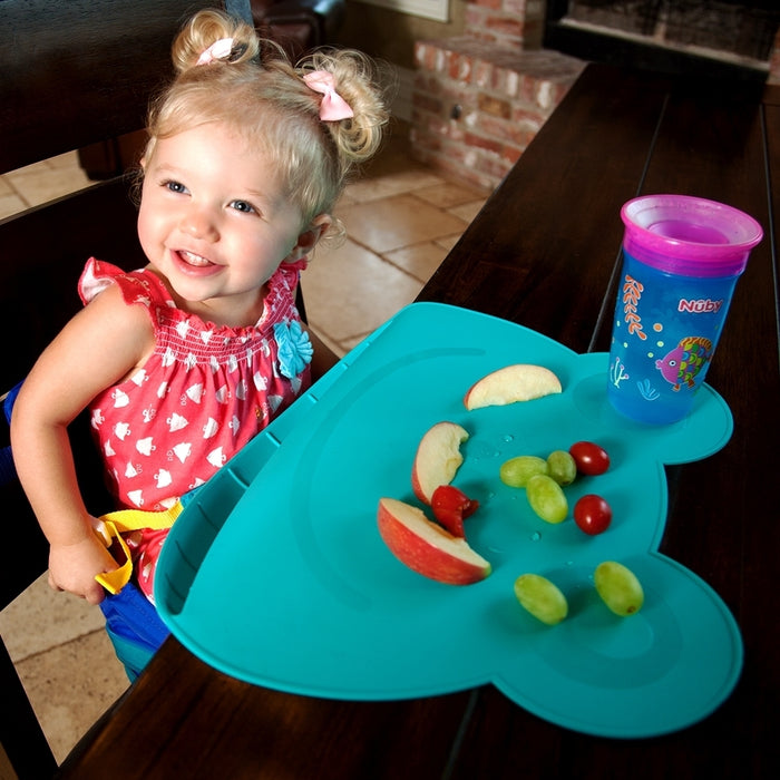 A smiling toddler with blonde pigtails sits at a table in a red dress with white hearts. She eats apple slices, grapes, and tomatoes on the Nuby Sure Grip Miracle Mat with Food-Catching Pouch, with a blue-purple cup beside her.