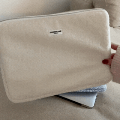A hand with red nails holds a beige Teddy Dolly laptop sleeve over a silver laptop on a beige sofa.