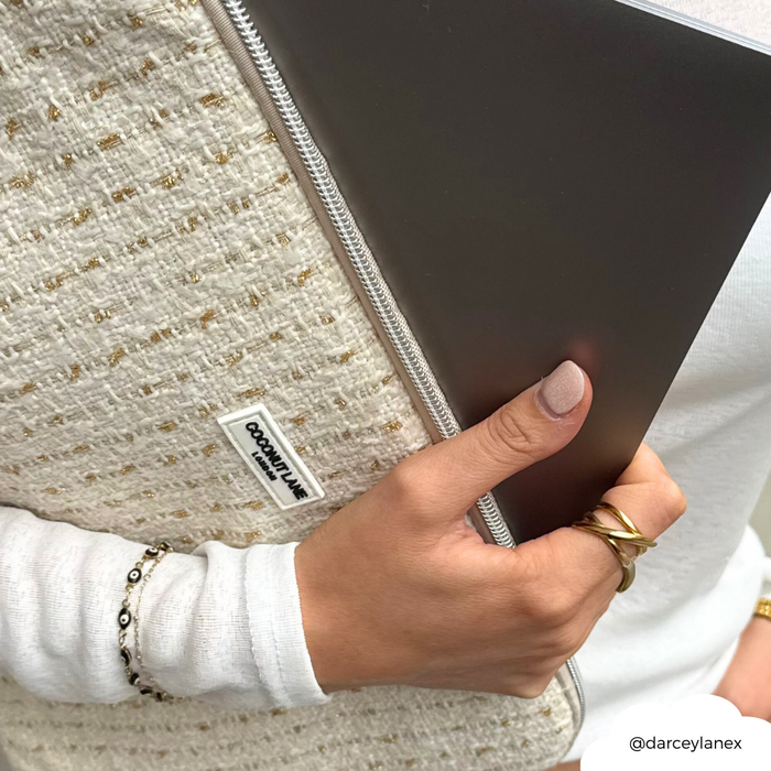 Gray laptop in a vanilla textured tweed sleeve, held by a hand with gold rings and a beaded bracelet.